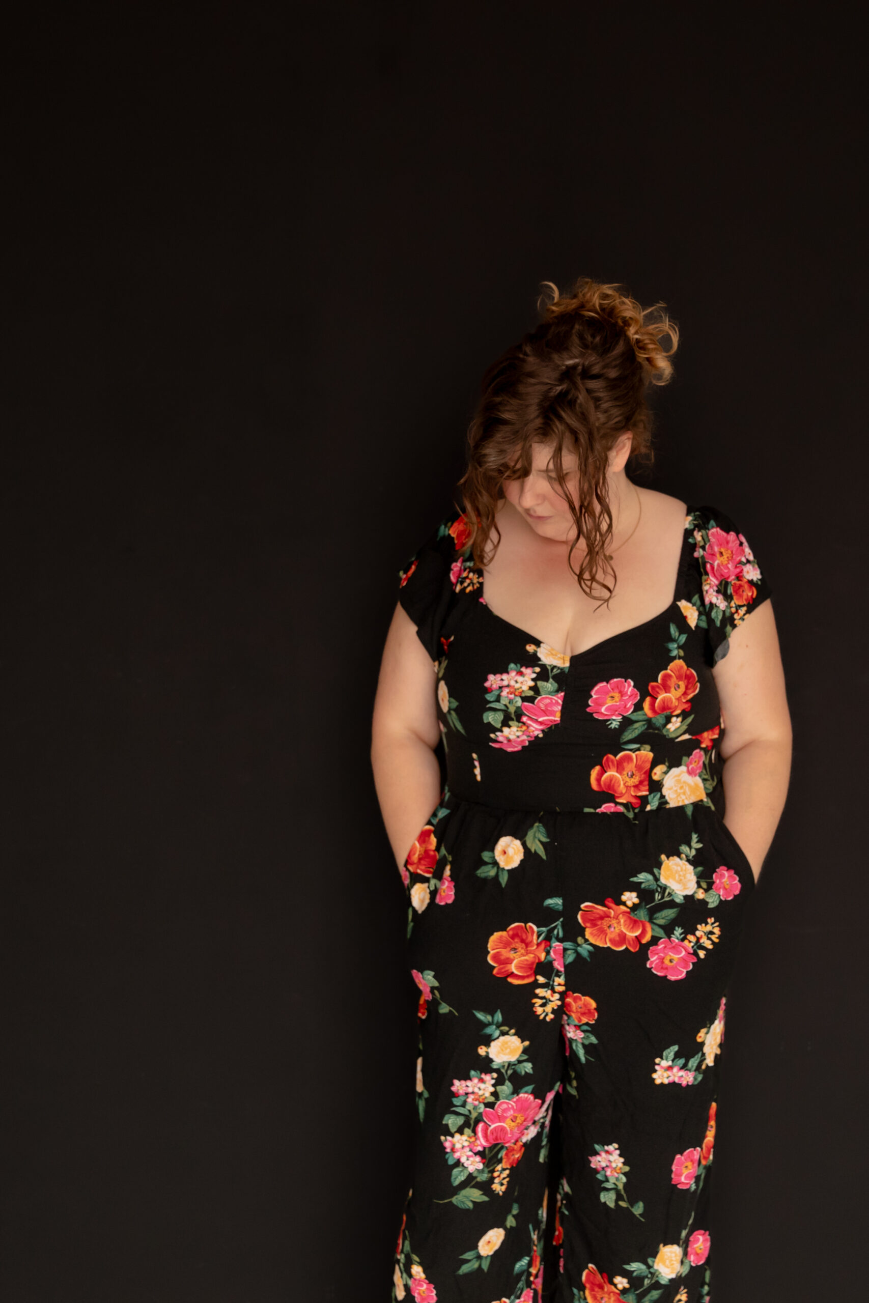 woman stands agains black wall in floral jumpsuit, looks down to the ground at studiostudio in ann arbor michigan