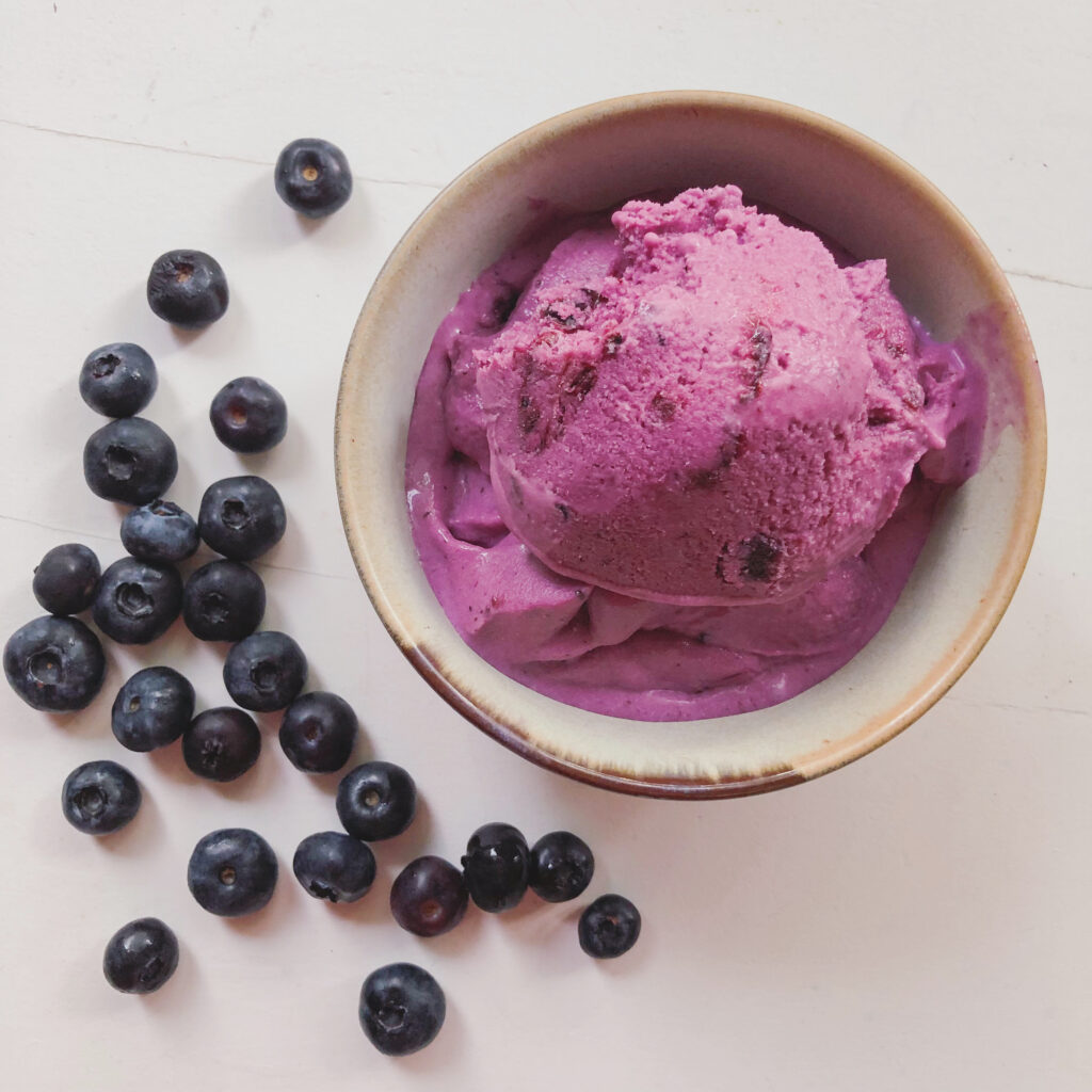 homemade blueberry cardamom ice cream in a small bowl on white table with blueberries sprinkled around