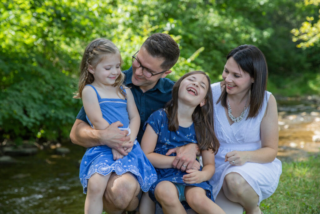 family of four cuddles and laughs near river at Mill Pond Park in Saline Michigan