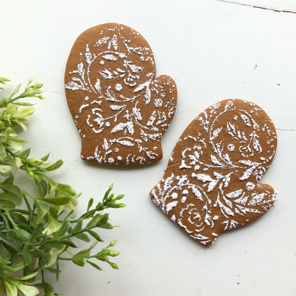 gingerbread mittens with ornate floral powdered sugar sprinkled on top next to greenery on white table