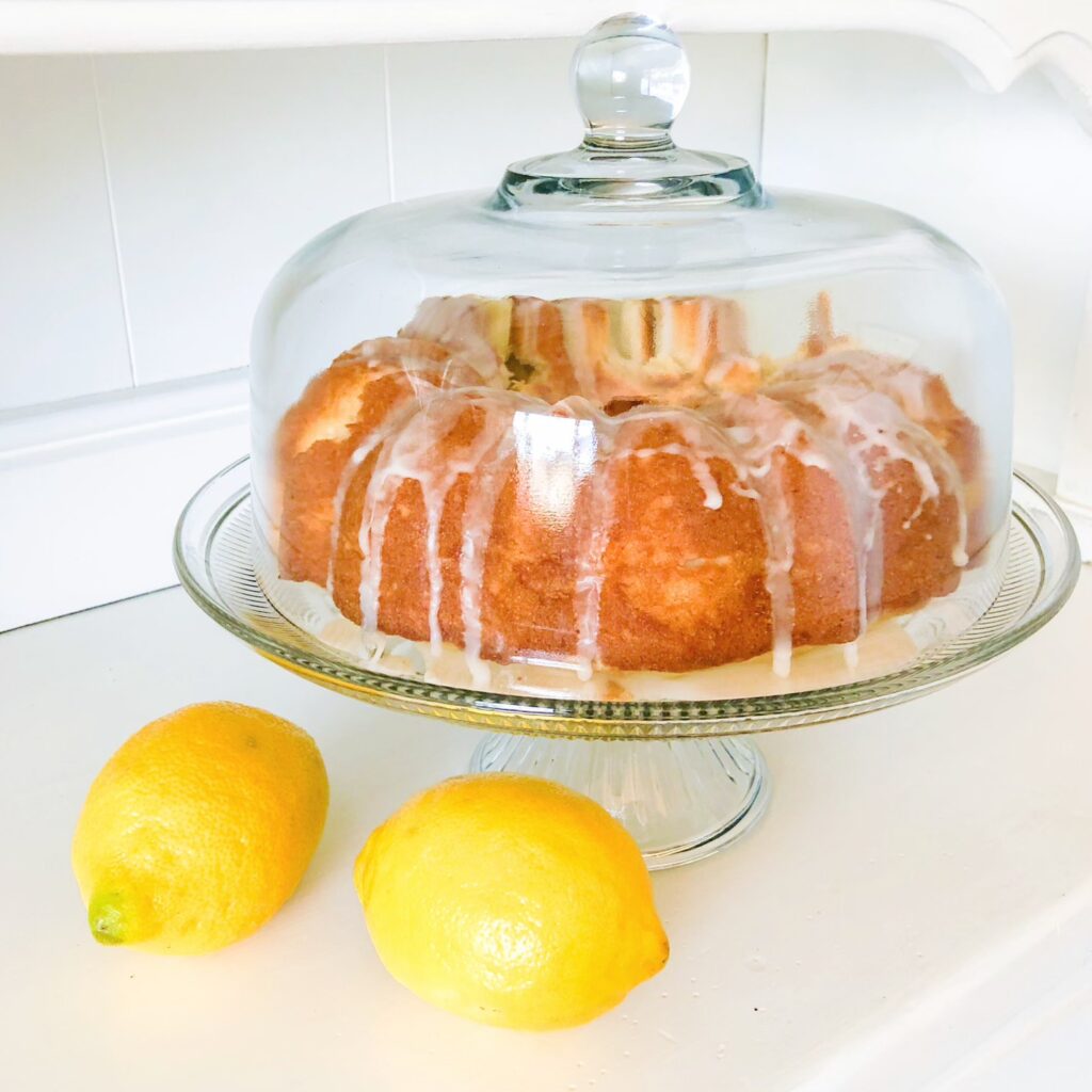 lemon bundt cake on glass cake stand with cover sits on white table top with two lemons