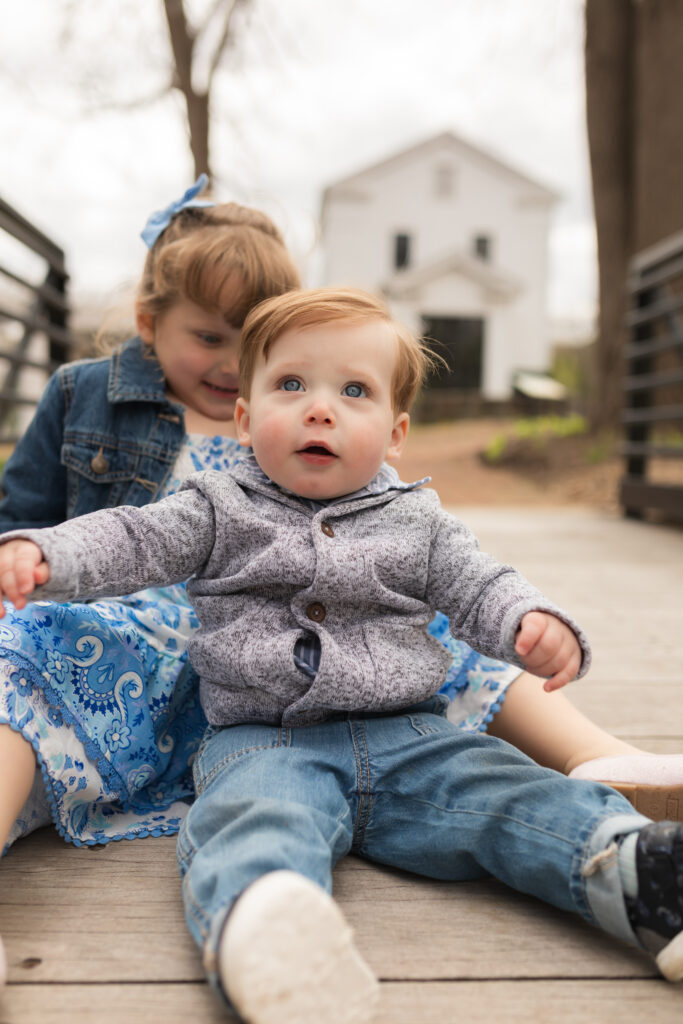 Baby boy and big sister sit on bridge with mill in background at Sharon Mills County Park in Manchester Michigan