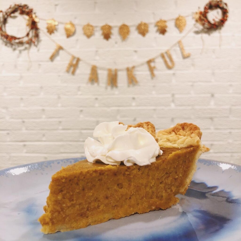 slice of pumpkin pie with small dollop of whipped cream on top on blue plate with thanksgiving decorations in the background that say thankful