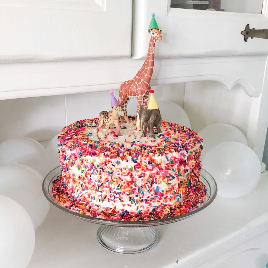 White cake with rainbow sprinkles and toy animals wearing tiny birthday hats with pom poms