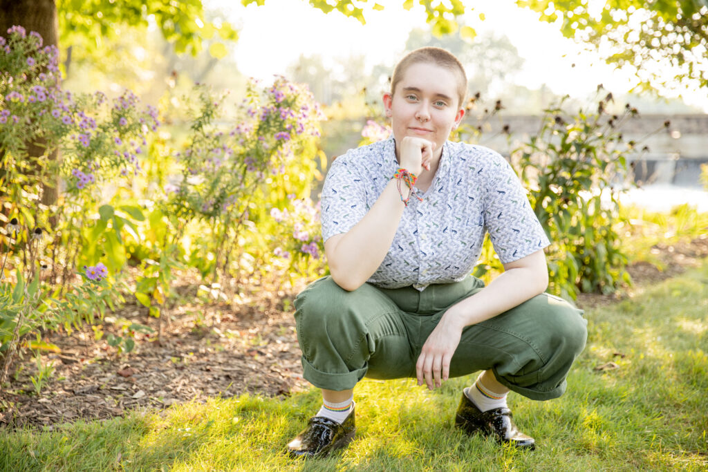 teenager poses for senior photos, squats in front of purple flowers and river with damn in the background during golden hour