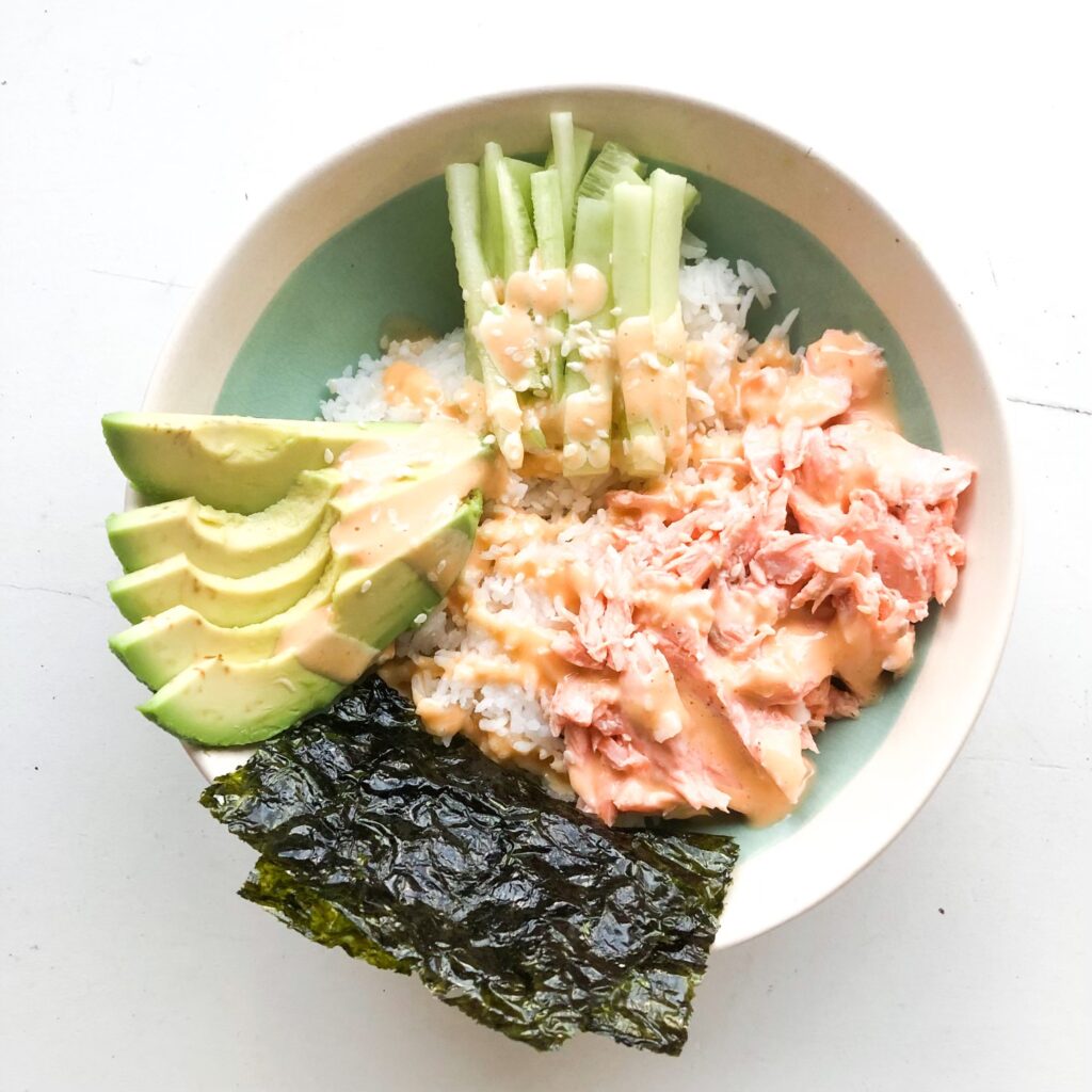 salmon sushi bowl with seaweed paper, sliced avocado and cucumber, on top of white rice with sesame seeds and yum yum sauce on top