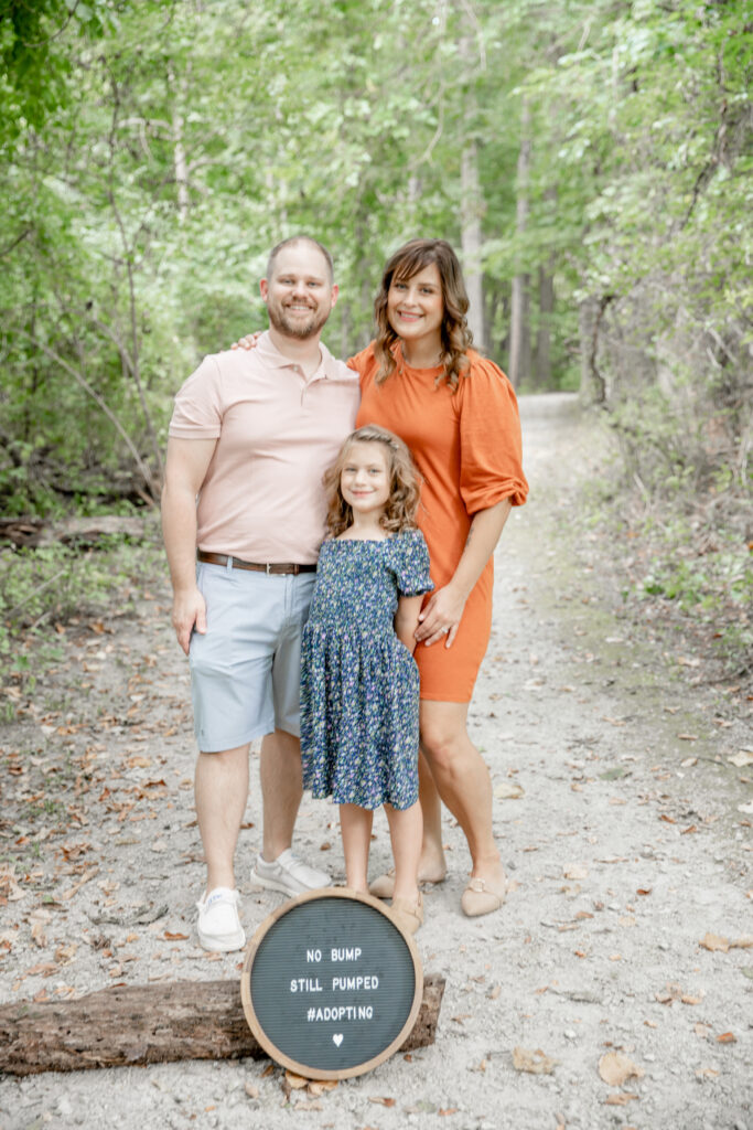 Family of three stands on a path with an adoption announcement sign in front of them