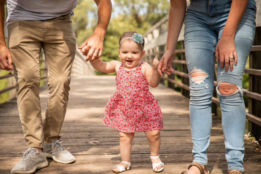 close up of one year old girl in red floral sun dress with blue flower headband walks across arched bridge holding mom and dad's hands and smiles at Gallup Park in Ann Arbor