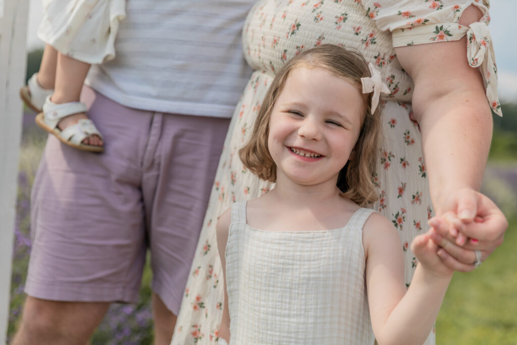 Toddler girls snuggles with mom and holds hand while smiling at camera at Lavender Lane in Milan Michigan