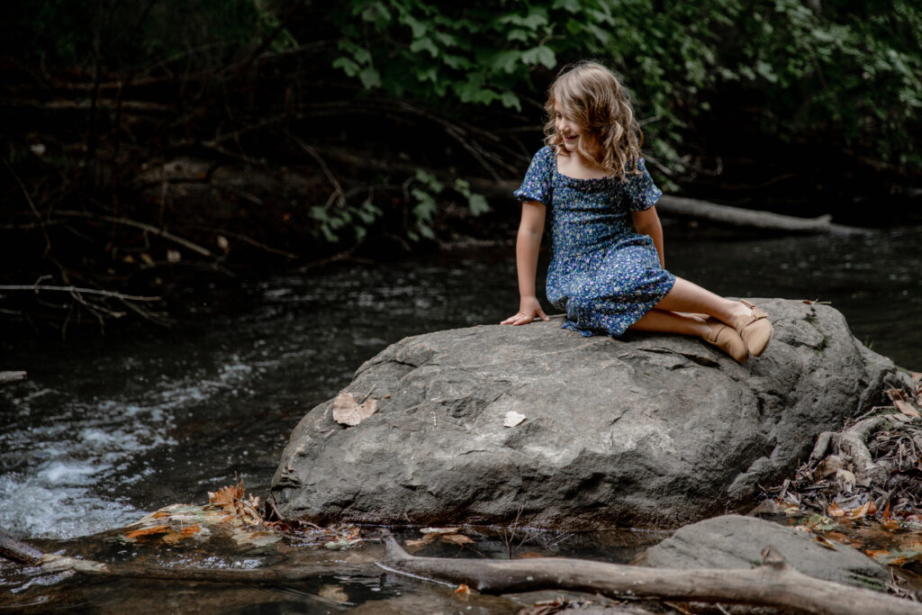 young girl sits on a rock on the edge of a river at Park Mill County Park in Ann arbor michigan