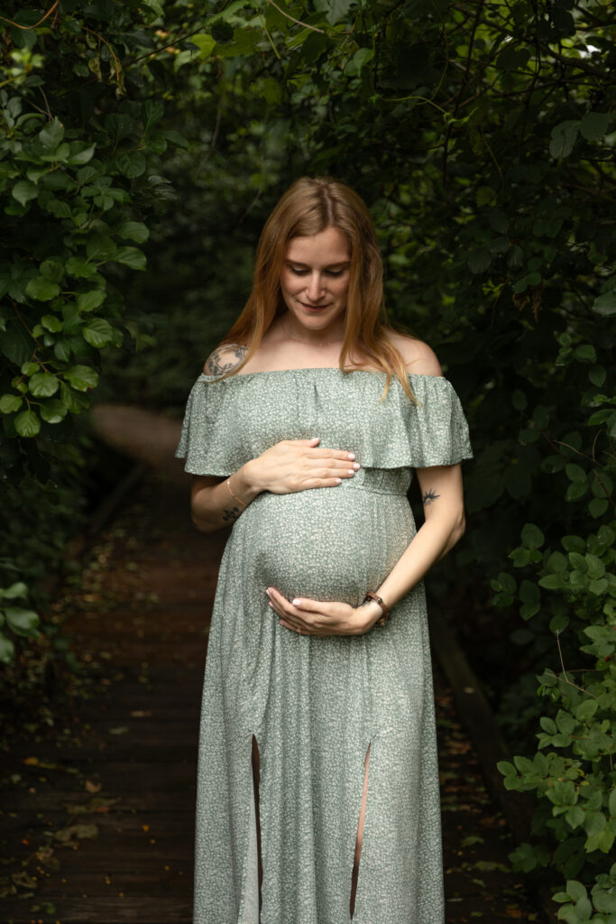 Pregnant woman stands and looks down at her belly while wrapping arms around it on a boardwalk in the woods at Saginaw Forest in Ann Arbor Michigan