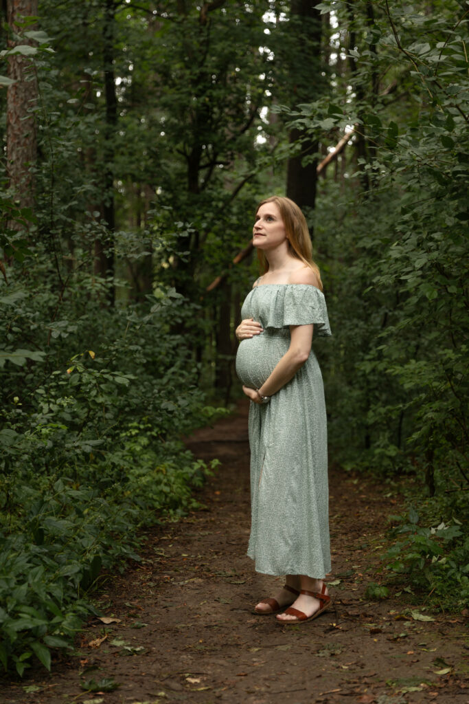 Woman stands on path looking up into the trees holding her pregnant belly at Saginaw Forest in Ann Arbor Michigan