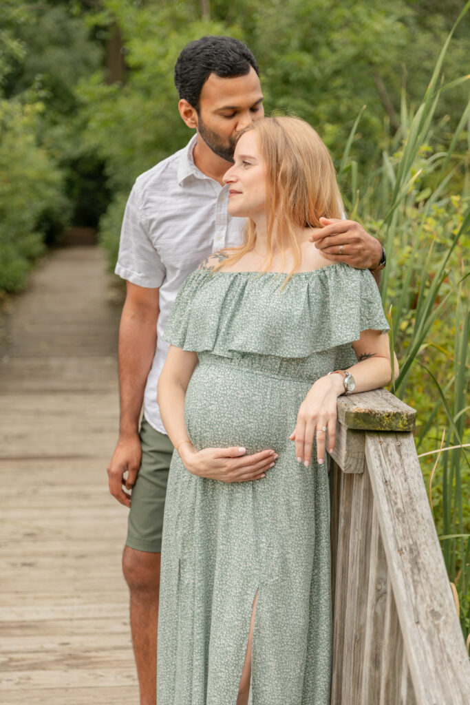 Soon to be father and mother stand on a boardwalk while she holds her belly and he kisses her head at Saginaw Forest in Ann Arbor Michigan