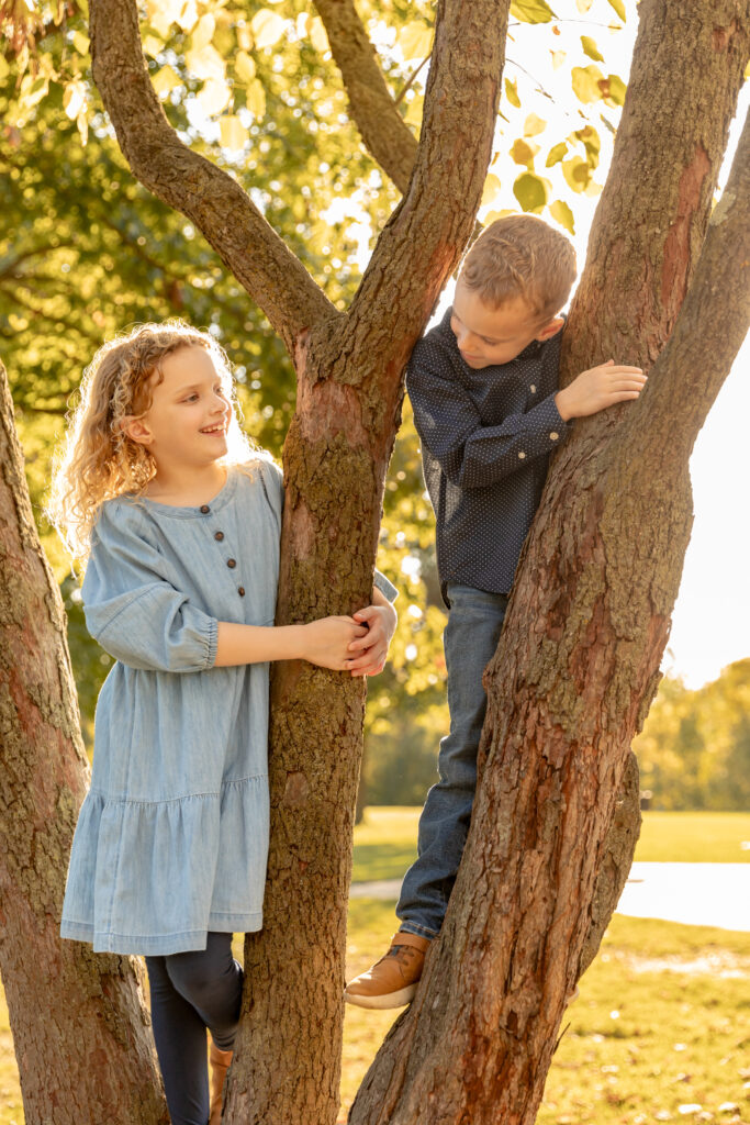 Brother and sister smile at each other while perched in tree at golden hour at Gallup Park in Ann Arbor Michigan