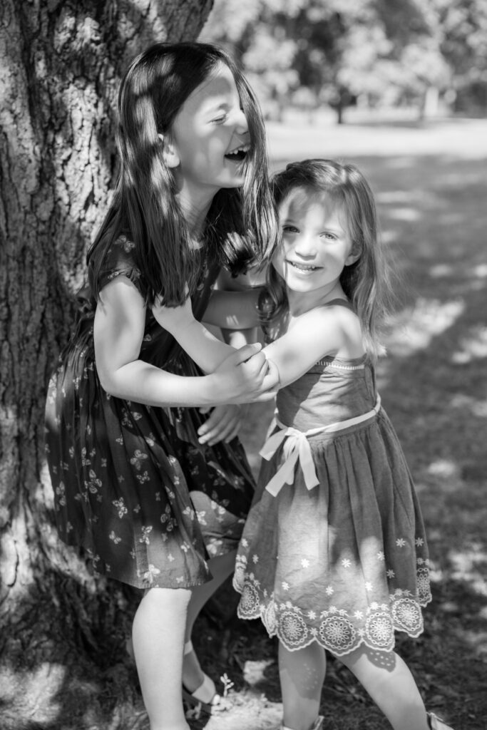 black and white image of young sisters laughing near tree at Mill Pond Park in Saline Michigan