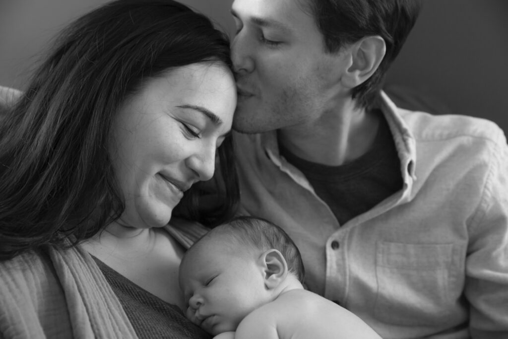 black and white photo of father kissing mother's head while she looks down at their sleeping newborn resting on her chest