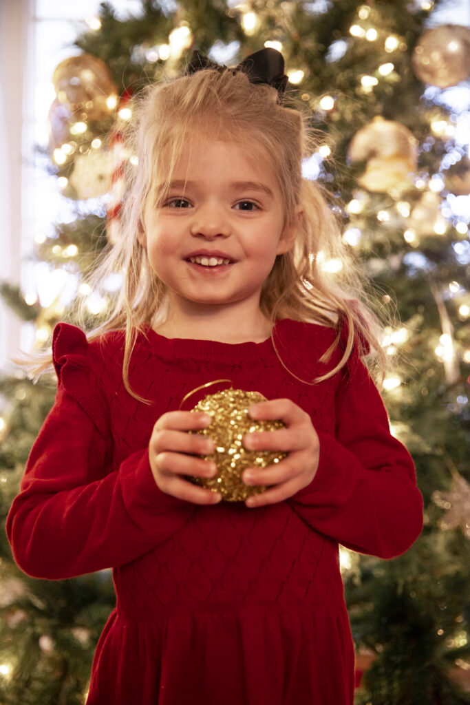 toddler girl holds golden christmas ornament while standing in front of christmas tree