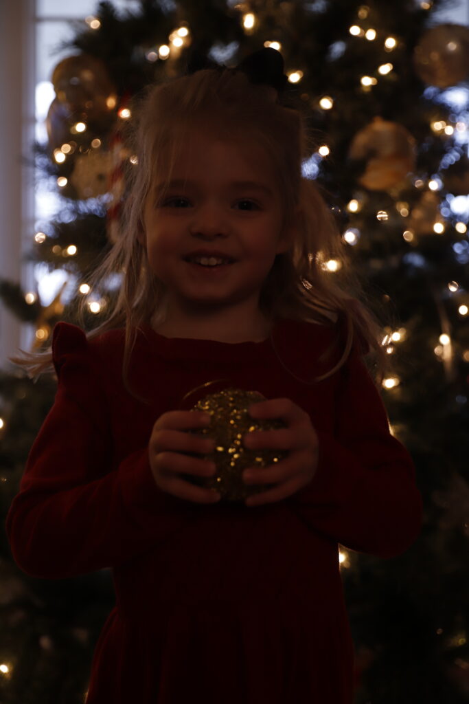 raw photo of toddler girl holding golden christmas ornament standing in front of christmas tree