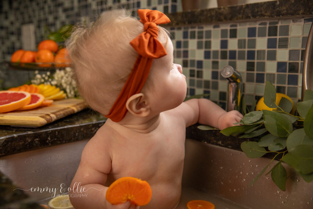 Baby girl sits in milk bath in the kitchen sink surrounded by sliced lemons, oranges, and grapefruits with baby's breath and eucalyptus