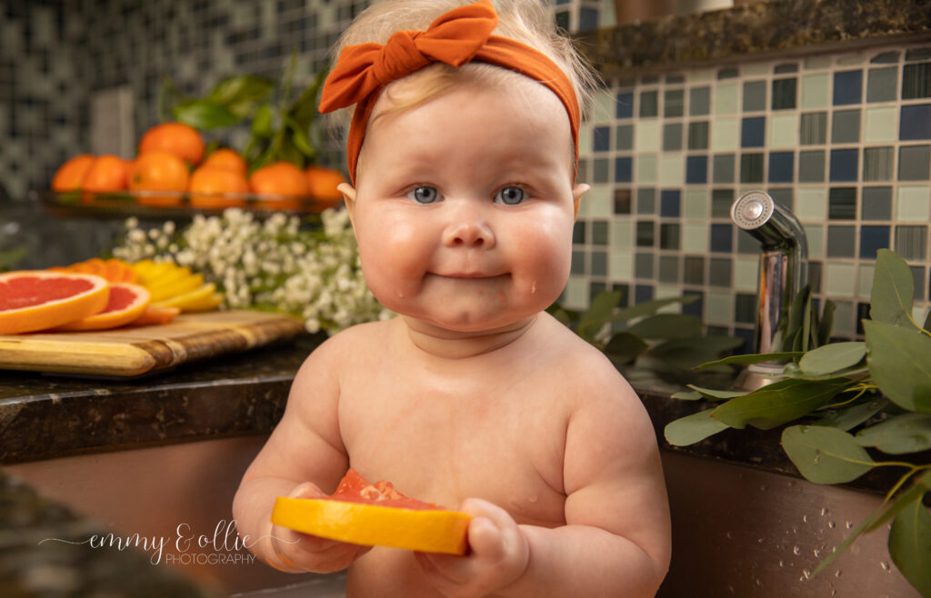 Baby girl sits in milk bath in the kitchen sink surrounded by sliced lemons, oranges, and grapefruits with baby's breath and eucalyptus