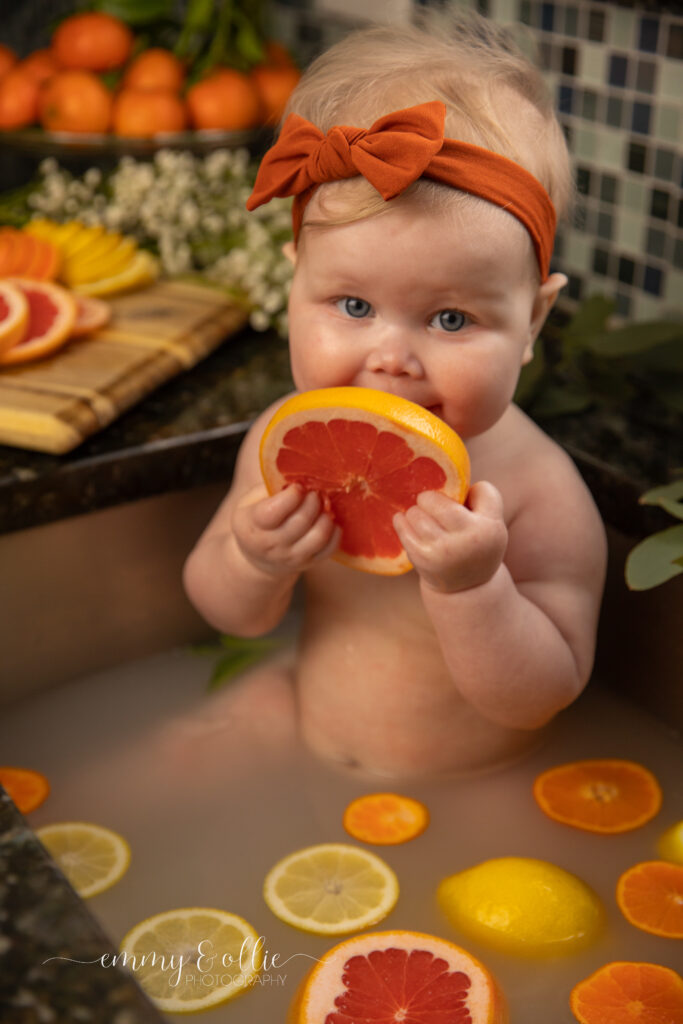 Baby girl sits in milk bath in the kitchen sink surrounded by sliced lemons, oranges, and grapefruits with baby's breath and eucalyptus