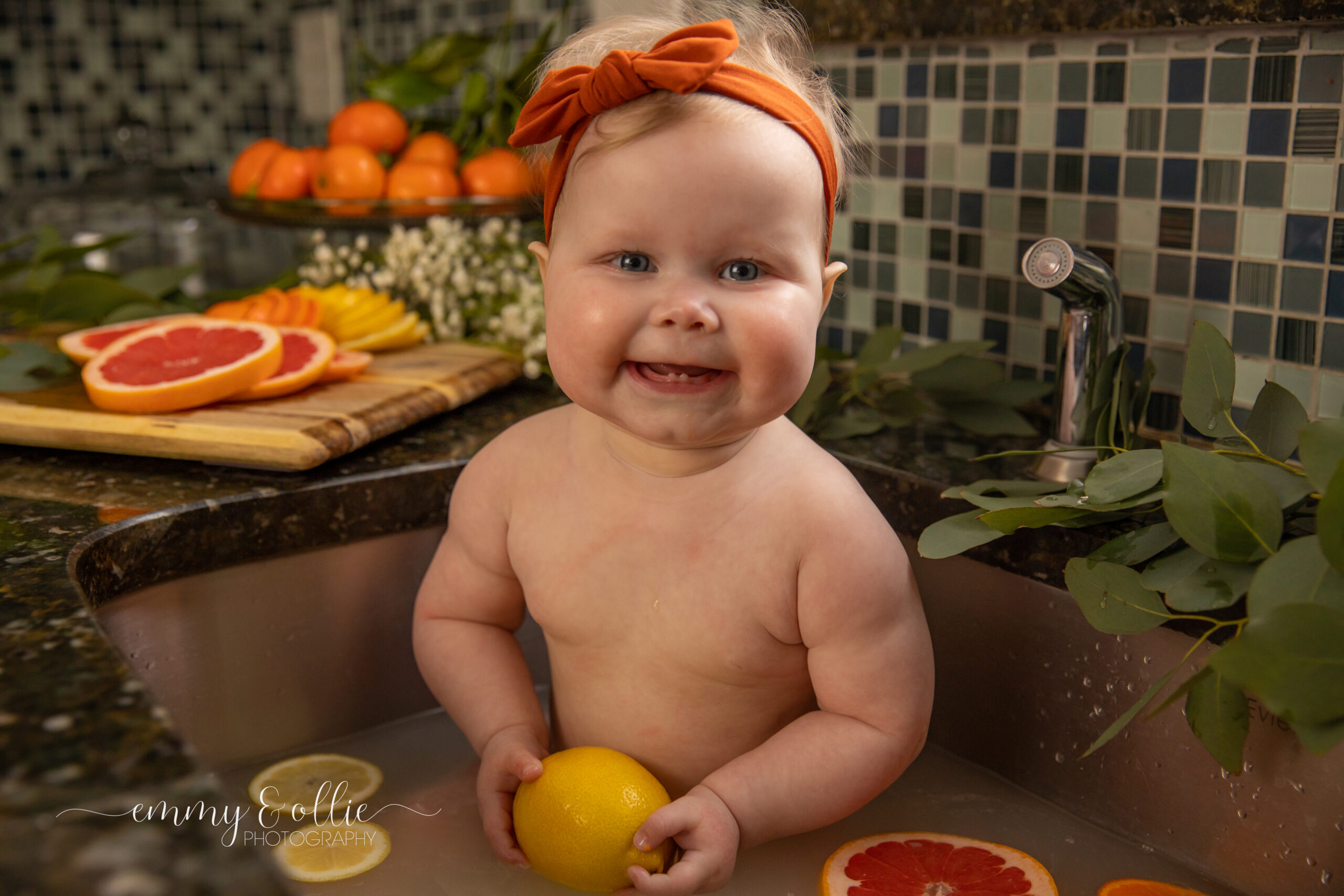 Baby girl sits in milk bath in the kitchen sink surrounded by sliced lemons, oranges, and grapefruits with baby's breath and eucalyptus