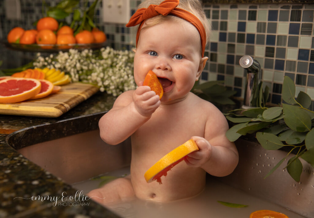 Baby girl sits in milk bath in the kitchen sink surrounded by sliced lemons, oranges, and grapefruits with baby's breath and eucalyptus