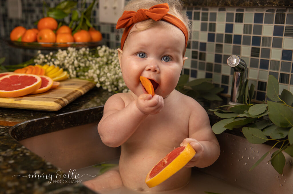 Baby girl sits in milk bath in the kitchen sink surrounded by sliced lemons, oranges, and grapefruits with baby's breath and eucalyptus