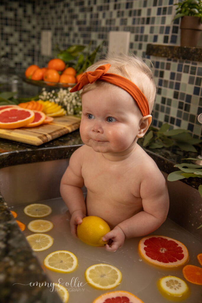 Baby girl sits in milk bath in the kitchen sink surrounded by sliced lemons, oranges, and grapefruits with baby's breath and eucalyptus