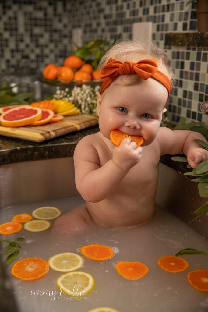 Baby girl sits in milk bath in the kitchen sink surrounded by sliced lemons, oranges, and grapefruits with baby's breath and eucalyptus