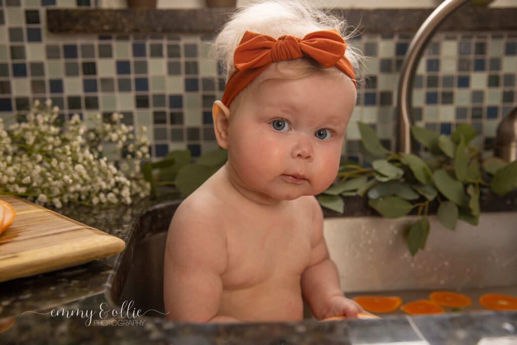 Baby girl sits in milk bath in the kitchen sink surrounded by sliced lemons, oranges, and grapefruits with baby's breath and eucalyptus