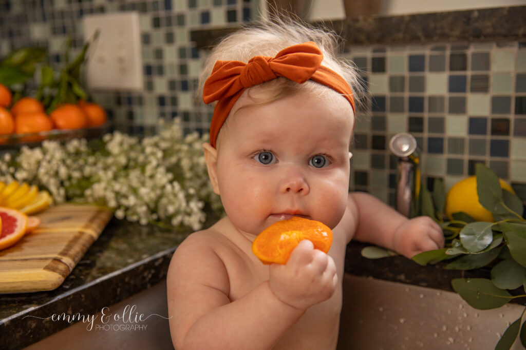 Baby girl sits in milk bath in the kitchen sink surrounded by sliced lemons, oranges, and grapefruits with baby's breath and eucalyptus