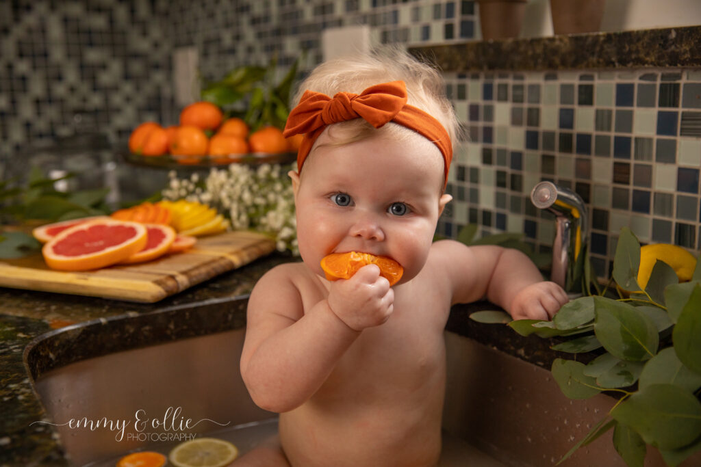 Baby girl sits in milk bath in the kitchen sink surrounded by sliced lemons, oranges, and grapefruits with baby's breath and eucalyptus