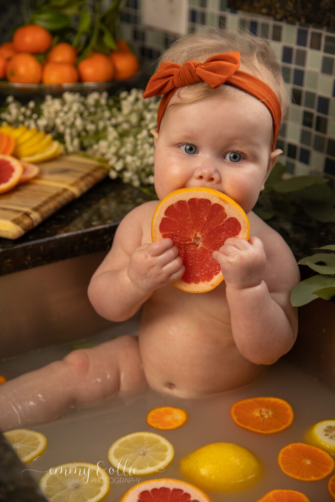 Baby girl sits in milk bath in the kitchen sink surrounded by sliced lemons, oranges, and grapefruits with baby's breath and eucalyptus