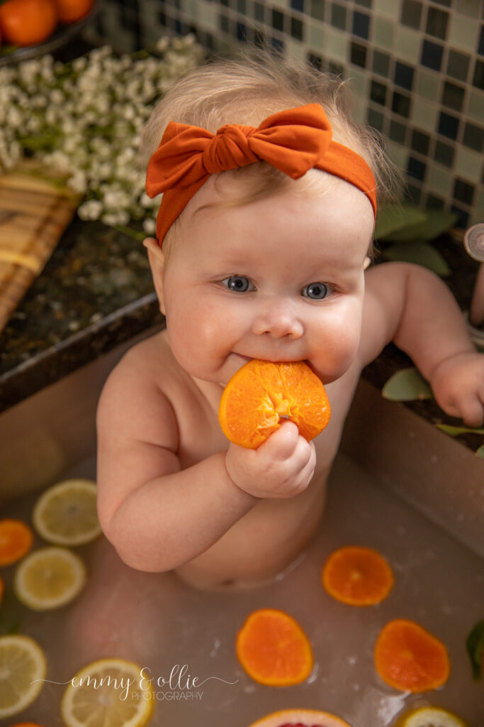 Baby girl sits in milk bath in the kitchen sink surrounded by sliced lemons, oranges, and grapefruits with baby's breath and eucalyptus