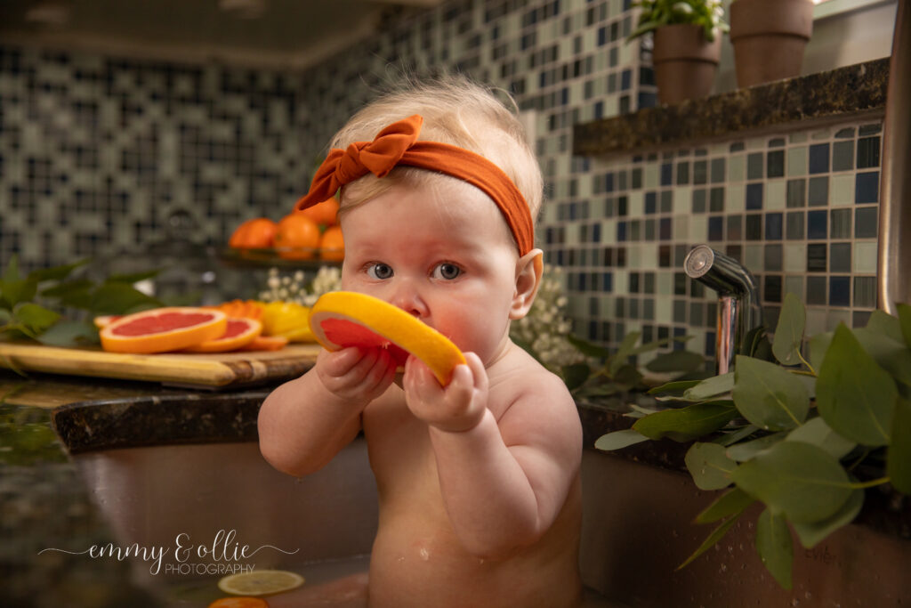 Baby girl sits in milk bath in the kitchen sink surrounded by sliced lemons, oranges, and grapefruits with baby's breath and eucalyptus