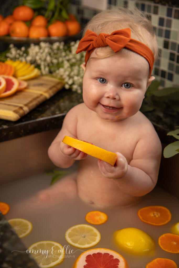 Baby girl sits in milk bath in the kitchen sink surrounded by sliced lemons, oranges, and grapefruits with baby's breath and eucalyptus