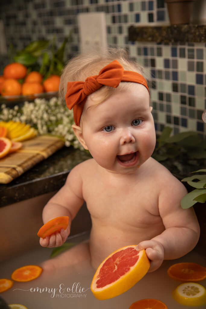 Baby girl sits in milk bath in the kitchen sink surrounded by sliced lemons, oranges, and grapefruits with baby's breath and eucalyptus