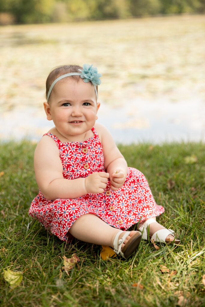 toddler girl sits on grass in front of pond and smiles at camera