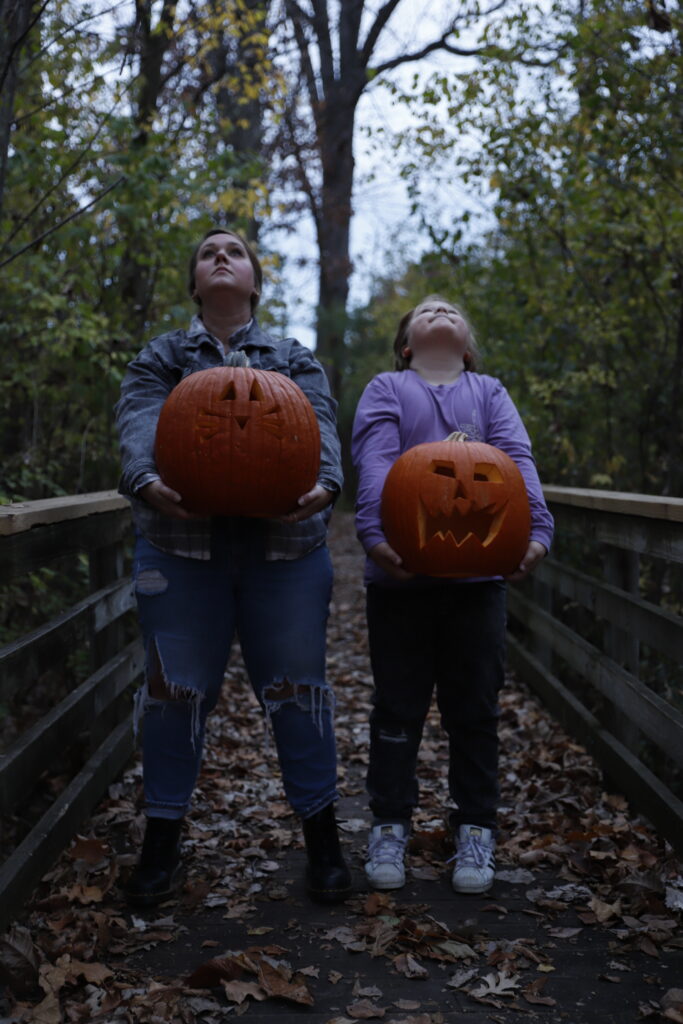 Raw photo of mother and son holding carved pumpkins in front of them on wooden bridge in the woods for halloween