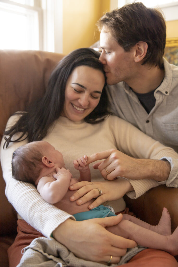 father leans over and kisses mother's head while newborn baby is cuddled in mother's arms, holding onto father's finger