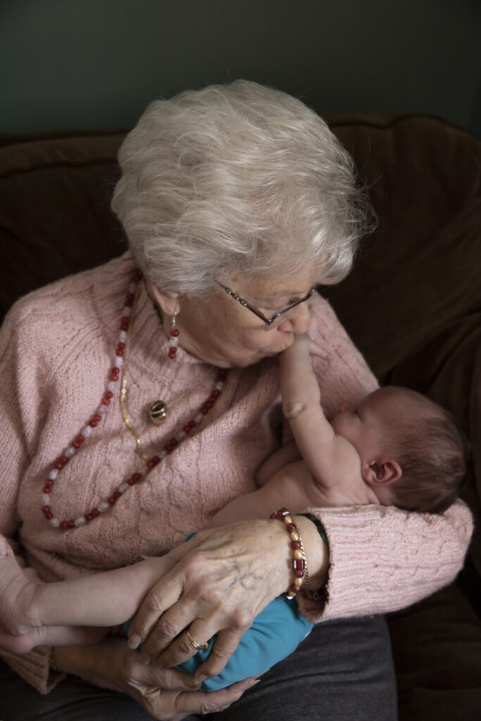 great grandmother kisses newborn great granddaughter on arm as she reaches up