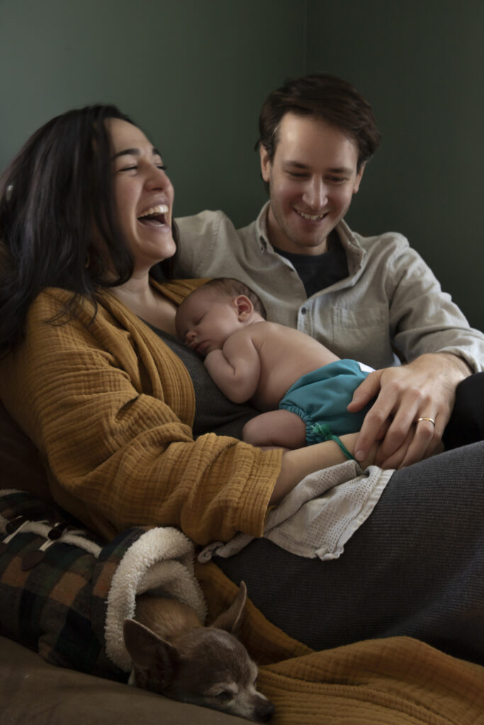 mother laughs while holding sleeping newborn baby on chest while father and chihuahua cuddle up with her