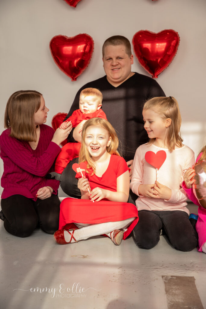 family sits on floor and laughs in front of white wall decorated with red heart balloons for valentine's day