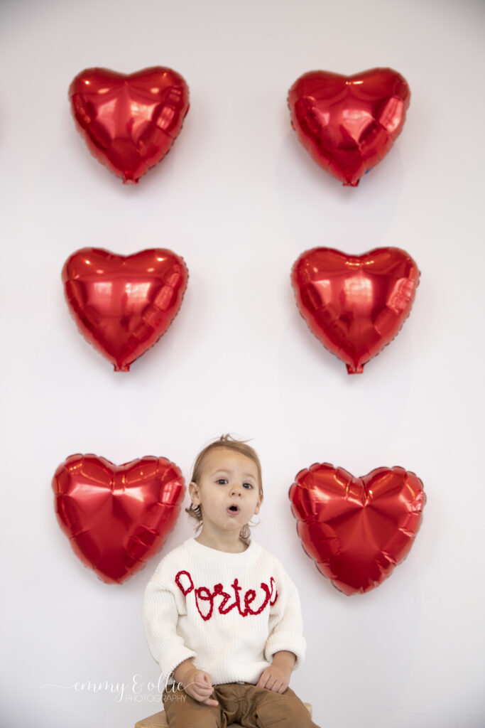 toddler boy sits in front of wall decorated with red heart balloons for valentines day