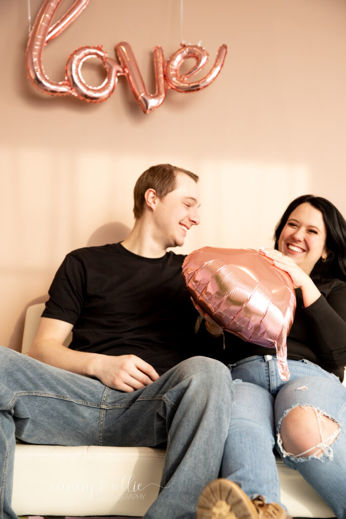 woman and man laugh together on a white couch with a pink heart balloon in front of pink wall decorated with pink love balloon