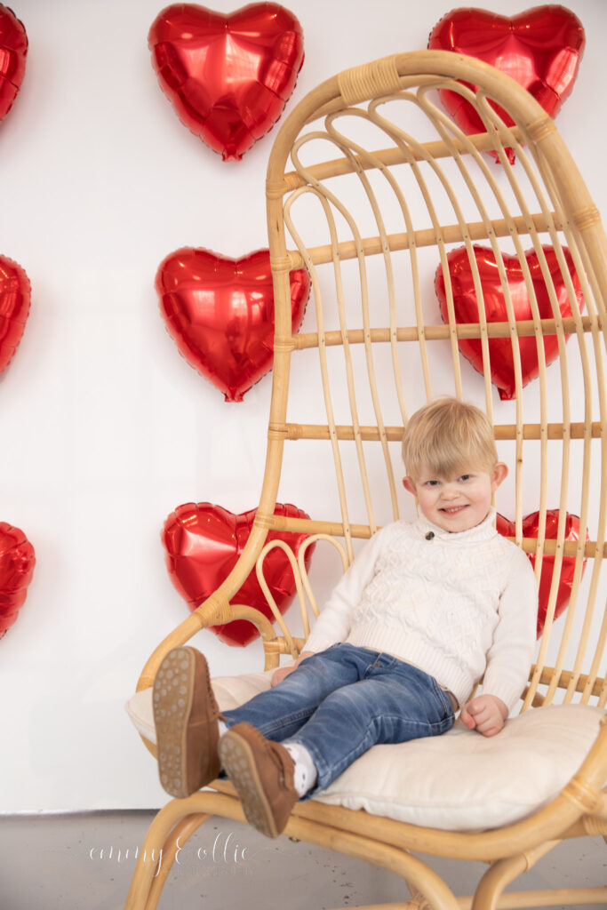 toddler boy sits in decorative wooden chair in front of white wall decorated with red heart balloons for valentine's day