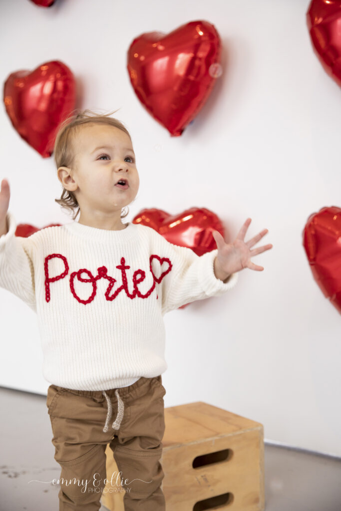 toddler boy stands and smiles at bubbles in front of wall decorated with red heart balloons for valentines day