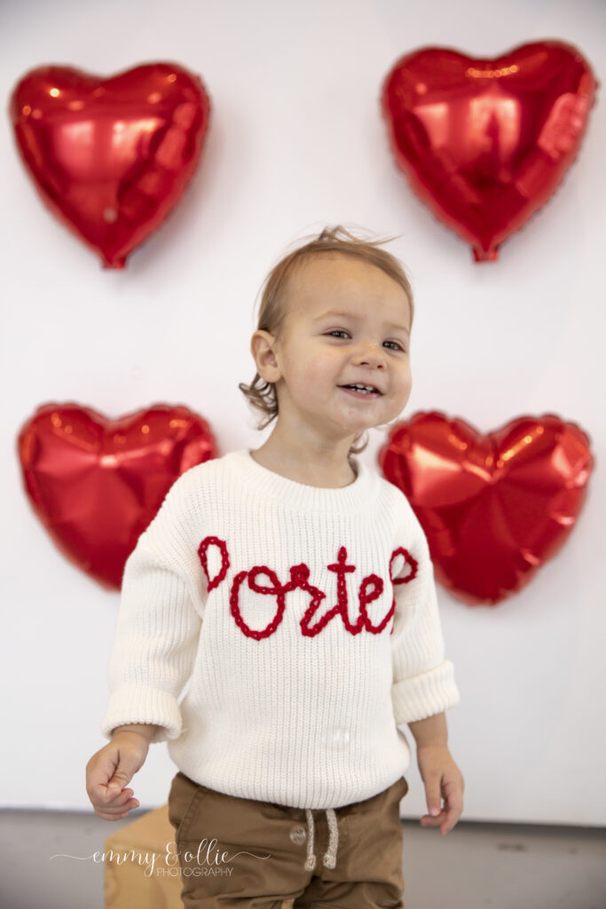 toddler boy stands and smiles in front of wall decorated with red heart balloons for valentines day