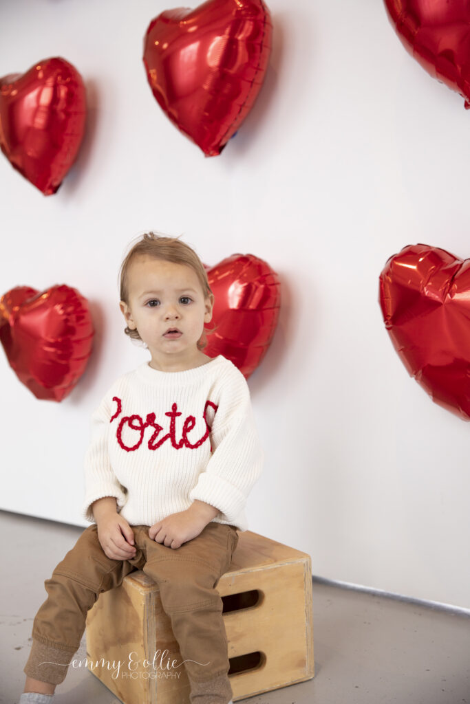 toddler boy sits in front of wall decorated with red heart balloons for valentines day